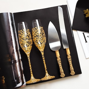 Könnte beinhalten: Zwei Sektgläser und ein Tortenset mit goldenem Filigranmuster. Das Set enthält einen Tortenheber und ein Messer. Das Set befindet sich auf einer schwarzen Oberfläche neben einer Zeitschrift.