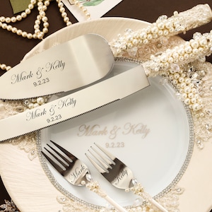 Puede incluir: Un juego de cuchillo y pala para pastel en plata con mangos incrustados de perlas, junto a dos tenedores a juego. Las hojas y tenedores est&aacute;n grabados con el texto Mark & Kelly 9.2.23. Un plato blanco presenta el mismo texto y un borde de cristal.