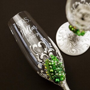 May include: A clear glass champagne flute featuring white scrollwork, green and white pearl embellishments, and the etched text "MRS. Hollar 5/23/26". A second matching glass with a white base and green pearl accents is partially visible nearby.