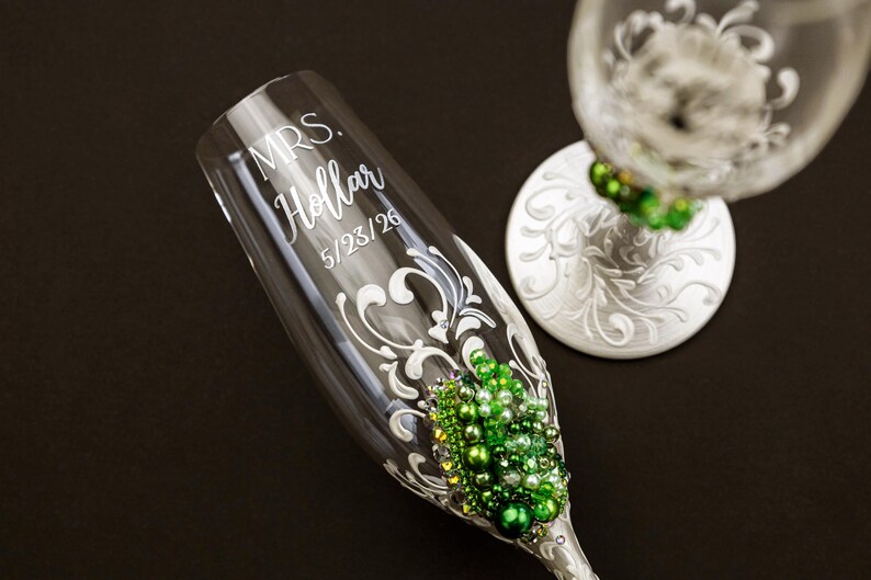 May include: A clear glass champagne flute featuring white scrollwork, green and white pearl embellishments, and the etched text "MRS. Hollar 5/23/26". A second matching glass with a white base and green pearl accents is partially visible nearby.