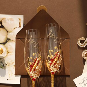 May include: Two clear glass champagne flutes featuring gold stems and intricate embellishments of red crystals, gold beads, and clear rhinestones. Each flute is etched with the letters J and Z in a cursive script, positioned over a dark brown envelope.