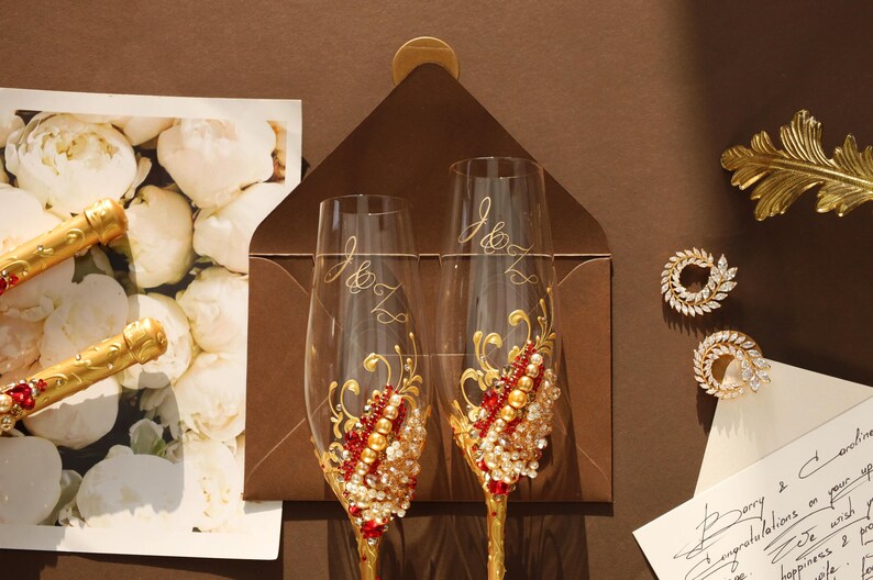 May include: Two clear glass champagne flutes featuring gold stems and intricate embellishments of red crystals, gold beads, and clear rhinestones. Each flute is etched with the letters J and Z in a cursive script, positioned over a dark brown envelope.