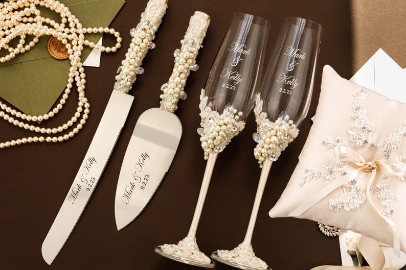 Puede incluir: Un juego de cuchillo y pala para pastel con mangos incrustados de perlas, dos copas de champ&aacute;n de cristal transparente con detalles de perlas y cristales, y un coj&iacute;n para anillos color crema con encaje. Cada art&iacute;culo tiene grabado Mark & Kelly 9.2.23.
