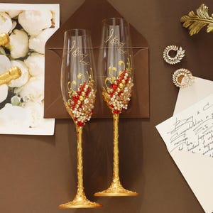 May include: Two clear glass champagne flutes with gold-painted stems and bases, featuring intricate gold scrollwork and clusters of red and white faux pearls and crystals. The glasses are etched with the initials J&Z and are decorated with ornate details.