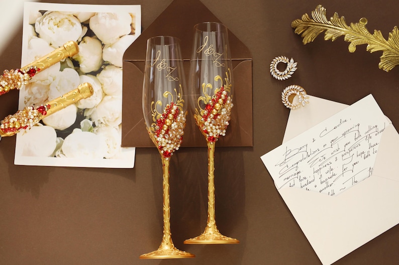 May include: Two clear glass champagne flutes with gold-painted stems and bases, featuring intricate gold scrollwork and clusters of red and white faux pearls and crystals. The glasses are etched with the initials J&Z and are decorated with ornate details.