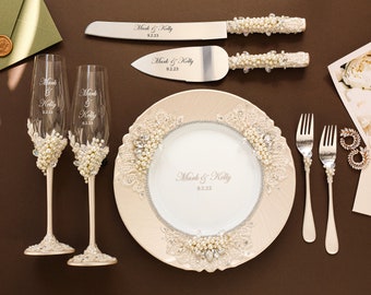 May include: A wedding cake cutting set with pearl-embellished handles, two champagne flutes with pearl accents, and two pearl-embellished forks. The set is on a beige charger plate with a white center and pearl accents. The plate has the names "Mark & Kelly" and the date "9.2.23" engraved on it.