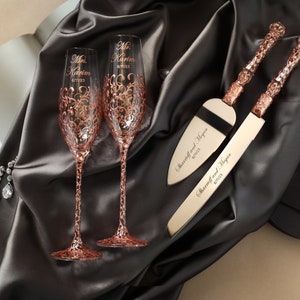 May include: A rose gold wedding champagne flute and cake serving set. The flutes are engraved with names and a date, and feature ornate detailing. The cake server and knife are also rose gold with matching engravings. The set is displayed on a satin cloth.