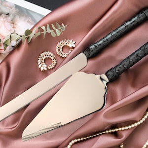 May include: A cake knife and server set with black ornate handles and silver blades, displayed on a mauve satin cloth. The set is accompanied by gold-toned jewelry and a string of pearls, suggesting a wedding or special occasion theme.