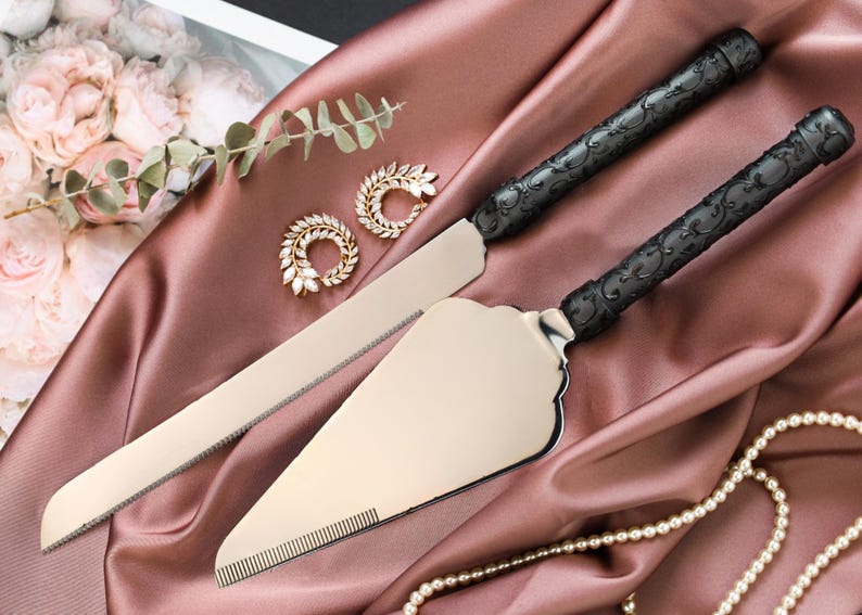 May include: A cake knife and server set with black ornate handles and silver blades, displayed on a mauve satin cloth. The set is accompanied by gold-toned jewelry and a string of pearls, suggesting a wedding or special occasion theme.