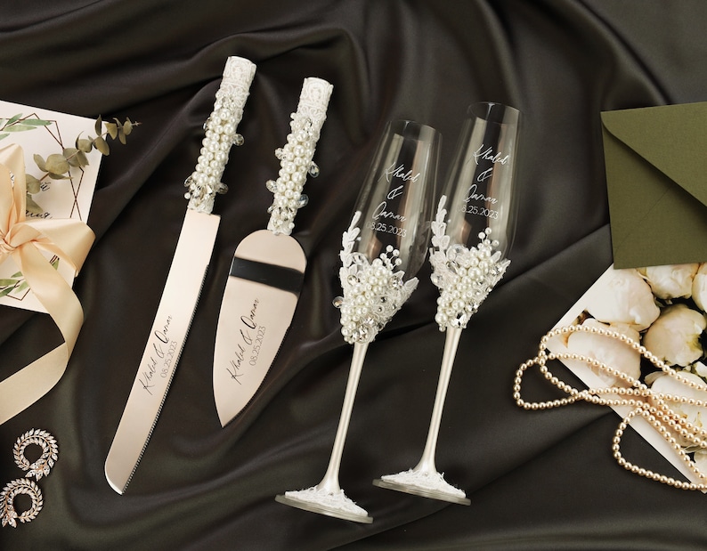 May include: A wedding cake cutting set with two silver knives and two champagne flutes. The knives are decorated with white pearl accents and the flutes are decorated with white pearl accents and lace. The knives are engraved with the text "Khaled & Omar 08.25.2023" and the flutes are engraved with the text "Khaled & Omar 08.25.2023".