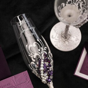 May include: A clear glass champagne flute featuring white scrollwork, purple and white beads, and the engraved text "Mr. Jording 9.12.26". A second glass with a white ornate stem and base is visible, alongside deep purple envelopes and stationery.