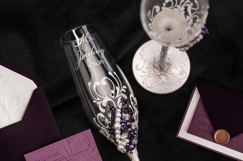 May include: A clear glass champagne flute featuring white scrollwork, purple and white beads, and the engraved text "Mr. Jording 9.12.26". A second glass with a white ornate stem and base is visible, alongside deep purple envelopes and stationery.