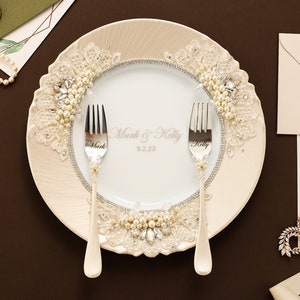 May include: A white ceramic plate featuring a cream-colored border with lace, pearls, and clear crystals. The center displays the text Mark & Kelly 9.2.23. Two silver forks are placed on the plate, each engraved with the names Mark and Kelly.