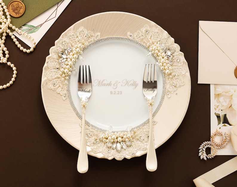 May include: A white ceramic plate featuring a cream-colored border with lace, pearls, and clear crystals. The center displays the text Mark & Kelly 9.2.23. Two silver forks are placed on the plate, each engraved with the names Mark and Kelly.