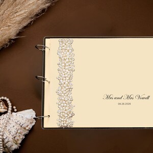 Puede incluir: Un libro de visitas de boda color crema con una columna vertebral con cuentas de perlas. El libro est&aacute; abierto, mostrando el texto "Mrs and Mrs Vowell" y la fecha "09.26.2026". Conchas marinas y perlas decorativas rodean el libro.