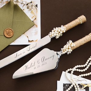 May include: A wedding cake knife and server set with pearl accents and twine-wrapped handles. The knife is engraved with "Love is Sweet", and the server with a name and date. A green envelope with a wax seal is in the background.