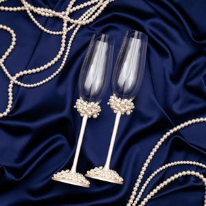 Puede incluir: Dos copas de champ&aacute;n con detalles de perlas descansan sobre una tela de sat&eacute;n azul oscuro, rodeadas de collares de perlas. Las copas tienen tallos blancos y racimos de perlas en la base y el borde.