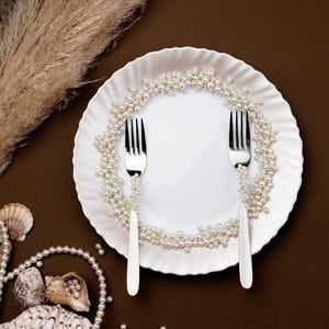 May include: A white plate with a ruffled edge is decorated with a pearl garland. Two silver forks are placed on the plate. Seashells and additional pearls are scattered around the plate on a brown background. Dried ornamental grass is in the background.