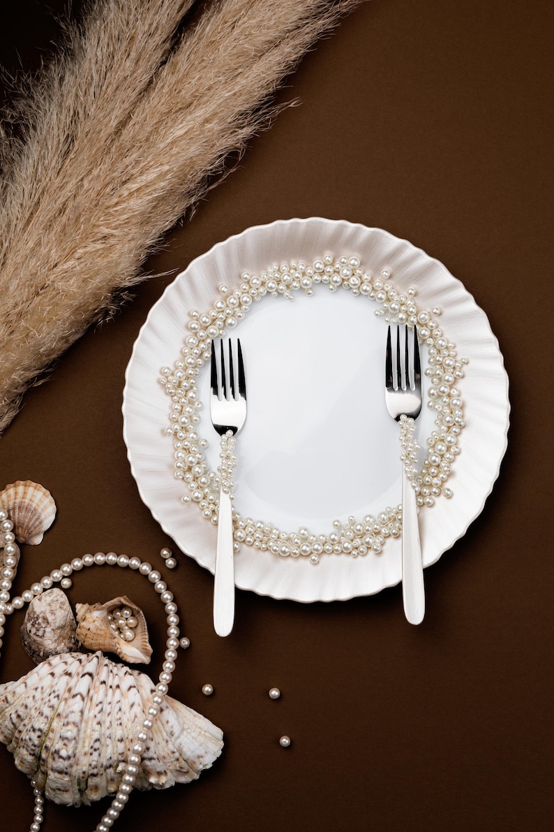 May include: A white plate with a ruffled edge is decorated with a pearl garland. Two silver forks are placed on the plate. Seashells and additional pearls are scattered around the plate on a brown background. Dried ornamental grass is in the background.