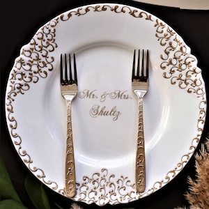 Puede incluir: Plato de cena de cer&aacute;mica blanca con un borde decorativo dorado y el texto "Mr. & Mrs. Shultz" en letras doradas. Dos tenedores de plata est&aacute;n colocados en el plato.