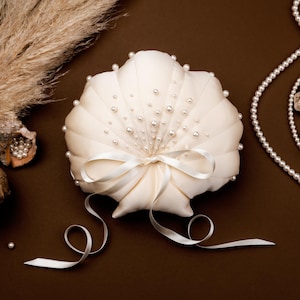 May include: A cream-colored, shell-shaped ring pillow adorned with small pearls and a satin bow. The pillow is surrounded by seashells, pearls, and dried plants, all set against a brown background. A decorative item for a wedding.