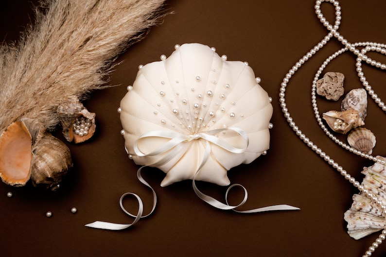May include: A cream-colored, shell-shaped ring pillow adorned with small pearls and a satin bow. The pillow is surrounded by seashells, pearls, and dried plants, all set against a brown background. A decorative item for a wedding.