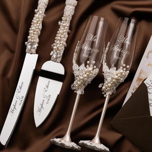 May include: A cake knife and server set with handles adorned in white lace, pearls, and crystals, alongside two champagne flutes featuring similar embellishments. The knife and server are engraved with Meghan & Ethan 11.14.26, while the flutes are etched with Mr. Stringer and Mrs. Stringer 11.14.26.