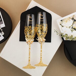 May include: Two champagne flutes with gold filigree and pearl accents, inscribed with "Mrs. Bond 6/23". The flutes are displayed with wedding photos, a card with white flowers, and a black envelope, set against a neutral background.