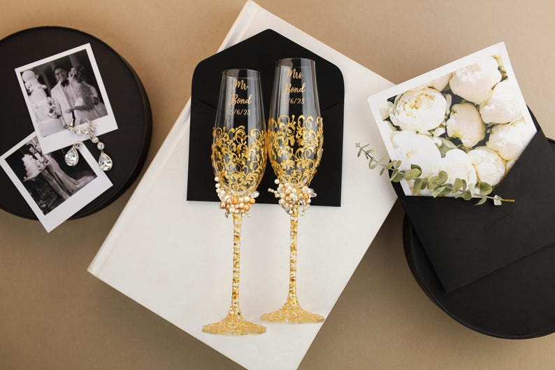 May include: Two champagne flutes with gold filigree and pearl accents, inscribed with "Mrs. Bond 6/23". The flutes are displayed with wedding photos, a card with white flowers, and a black envelope, set against a neutral background.