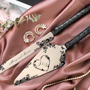 May include: A wedding cake knife and server set with black handles and decorative floral and skull accents. The server features a heart design with initials. The knife reads "Always and Forever". The set is displayed on a pink satin cloth.