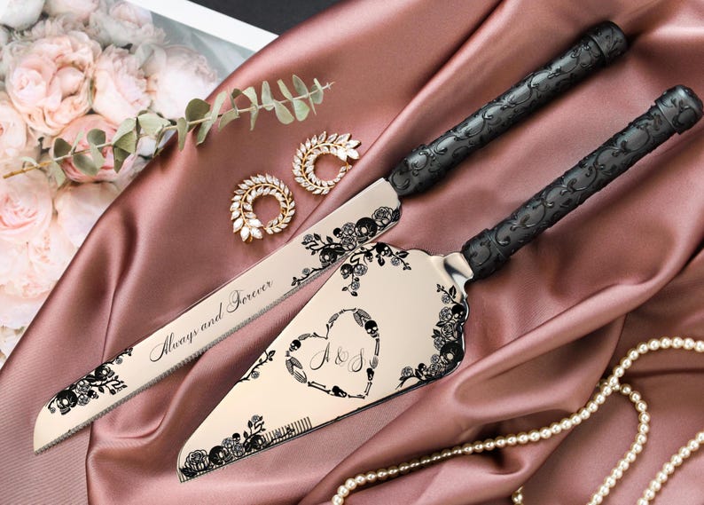 May include: A wedding cake knife and server set with black handles and decorative floral and skull accents. The server features a heart design with initials. The knife reads "Always and Forever". The set is displayed on a pink satin cloth.
