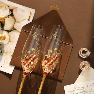 May include: Two clear glass champagne flutes featuring gold stems and intricate embellishments of red crystals, white pearls, and gold swirls. The glasses are engraved with the letters J and Z. Also included are two circular crystal earrings.
