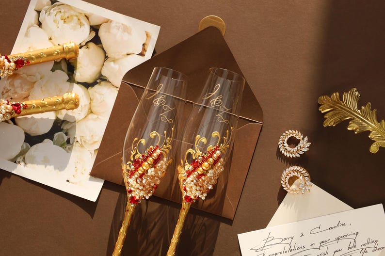 May include: Two clear glass champagne flutes featuring gold stems and intricate embellishments of red crystals, white pearls, and gold swirls. The glasses are engraved with the letters J and Z. Also included are two circular crystal earrings.