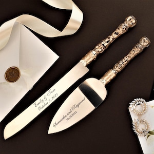 Könnte beinhalten: Ein Hochzeitstortenmesser- und Servier-Set mit verzierten goldenen Griffen und silbernen Klingen. Messer und Servierer sind mit Namen und Daten graviert. Ein weißer Umschlag mit goldenem Siegel und ein Paar Ohrringe sind ebenfalls im Bild.