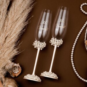 May include: Two champagne flutes with "Mr. & Mrs. Vow" script, adorned with pearl clusters. The stems are white, and the bases are also decorated with pearls. The image includes seashells, pearls, and dried plants, suggesting a wedding theme.