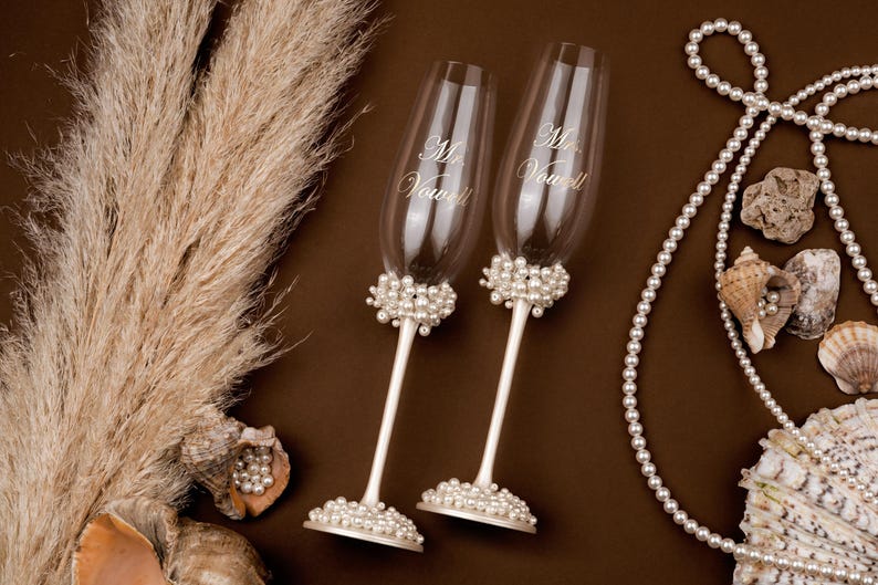 May include: Two champagne flutes with "Mr. & Mrs. Vow" script, adorned with pearl clusters. The stems are white, and the bases are also decorated with pearls. The image includes seashells, pearls, and dried plants, suggesting a wedding theme.