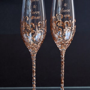 May include: Two champagne flutes with gold painted floral designs and the names "Terri & Seron" and the date "08.20.22" written on them.