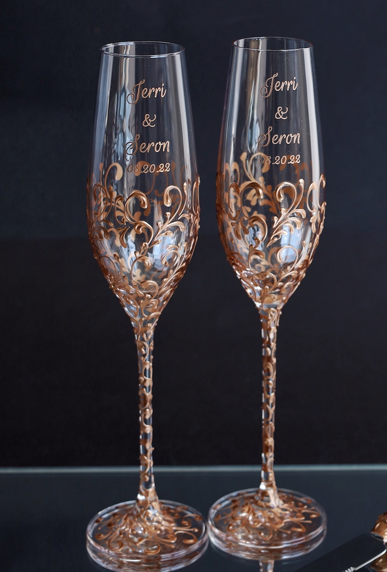 May include: Two champagne flutes with gold painted floral designs and the names "Terri & Seron" and the date "08.20.22" written on them.