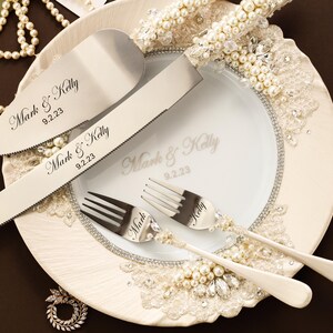 Puede incluir: Un juego de cuchillo y pala para pastel en plata con mangos incrustados de perlas, junto a dos tenedores de plata. Las piezas met&aacute;licas est&aacute;n grabadas con Mark & Kelly 9.2.23. Un plato blanco muestra el mismo texto, decorado con encaje y perlas.