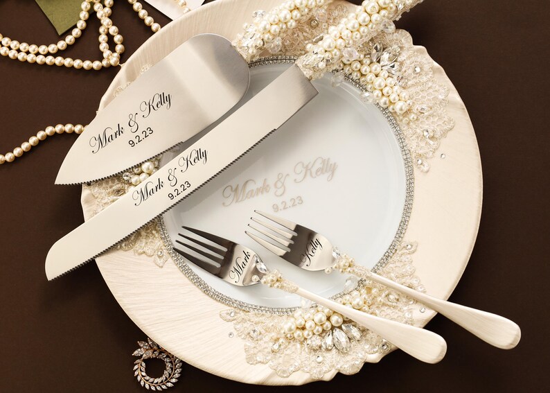 Puede incluir: Un juego de cuchillo y pala para pastel en plata con mangos incrustados de perlas, junto a dos tenedores de plata. Las piezas met&aacute;licas est&aacute;n grabadas con Mark & Kelly 9.2.23. Un plato blanco muestra el mismo texto, decorado con encaje y perlas.