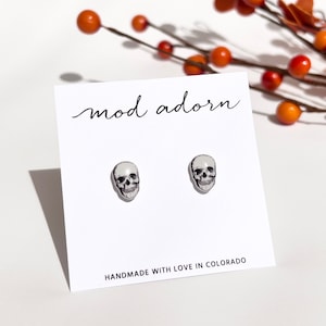 May include: A pair of silver skull stud earrings on a white card with the text "mod adorn" and "HANDMADE WITH LOVE IN COLORADO".