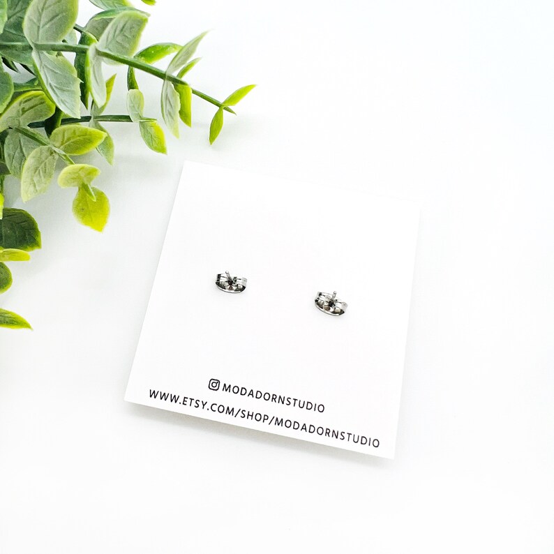 May include: A pair of silver-colored stud earrings on a white card. The card has the text "@MODADORNSSTUDIO" and "WWW.ETSY.COM/SHOP/MODADORNSSTUDIO". Green leaves are in the background.