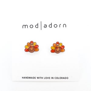 May include: A pair of small, colorful turkey stud earrings. The earrings are made of resin and feature a cartoon turkey with orange, yellow, and brown feathers. The earrings are handmade with love in Colorado.