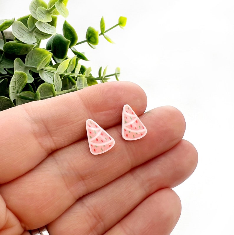 May include: A pair of Christmas tree-shaped earrings. The earrings are light pink with white frosting and red and green sprinkles. The earrings are small and triangular, with a festive design.
