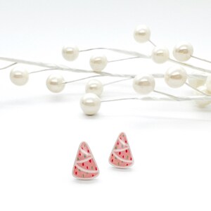 May include: Two Christmas tree-shaped stud earrings. The earrings are pink with white stripes and small green and red dots. The background is white with a decorative pearl-like branch.