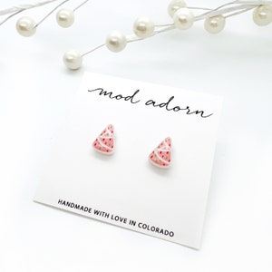 May include: A pair of watermelon slice earrings on a white card. The earrings are pink with white and red details. The card reads "mod adorn" and "HANDMADE WITH LOVE IN COLORADO".