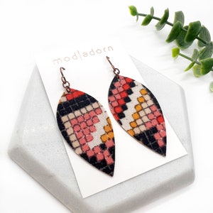 May include: A pair of dangle earrings with a geometric pattern in shades of red, black, white, and yellow. The earrings are shaped like a teardrop and have a copper-colored hook.