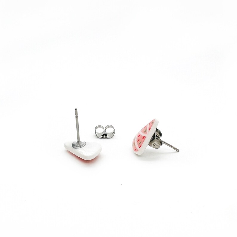 May include: White ceramic stud earrings with a red and pink geometric pattern. The earrings have stainless steel posts and butterfly backs. The earrings are displayed on a white background.