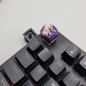 May include: A custom mechanical keyboard keycap featuring a small, sculpted white and orange cat sleeping on a square, textured purple cushion. The keycap is installed on a black keyboard near the Tab key, with a clear base visible underneath.