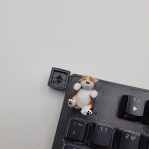 May include: A small, hand-painted corgi figurine attached to a black mechanical keyboard keycap. The dog has brown and white fur, black eyes, and visible paw pads. A separate black square keycap base sits next to the keyboard, which features keys labelled with Tab, Q, W, 1, 2, and F1.